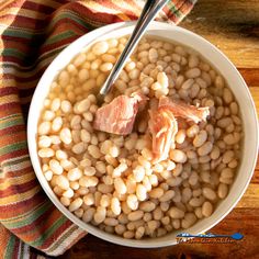 a white bowl filled with beans and meat on top of a wooden table next to a striped towel