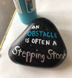 a black rock with writing on it next to a blue cup and marker board that says an obstacle is often a stepping stone