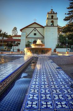 a fountain in front of a church with blue tiles on the ground and a clock tower behind it