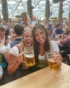 two women are smiling and holding beer glasses in front of them at an outdoor event