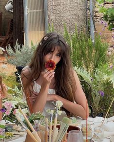 a woman sitting at a table with flowers in her hand and holding a flower up to her mouth