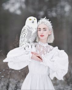 a woman wearing a white dress holding an owl on her shoulder in front of snow covered trees