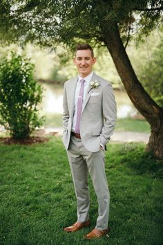 a man in a gray suit and pink tie standing under a tree with his hands in his pockets