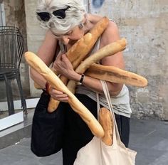 an older woman carrying several large baguettes in her hands while walking down the street