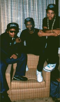 three young men sitting on a couch in front of a window with curtains behind them