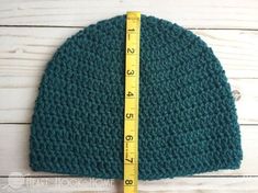 a green crocheted hat with a measuring tape on the side and a white wooden background