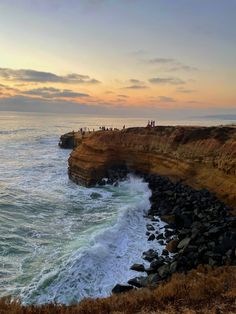 people are standing on the edge of a cliff overlooking the ocean and cliffs at sunset