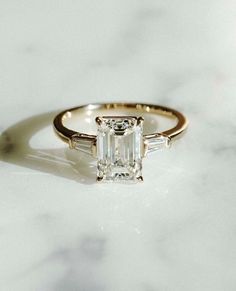 an emerald cut engagement ring with three baguets on the side, sitting on a marble surface