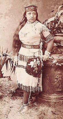 an old photo of a native american woman in traditional dress and headdress standing next to a planter