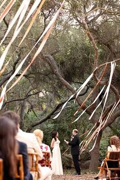 a couple getting married in the woods with streamers hanging from trees and people sitting on chairs