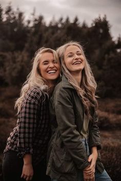 two young women standing next to each other in front of some trees and bushes with their arms around one another