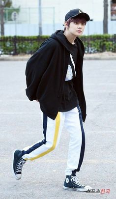 a young man in black jacket and white pants walking across a parking lot with his hat on