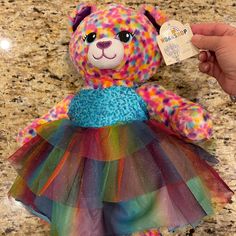 a colorful teddy bear wearing a multicolored tutu skirt with a tag in it's hand