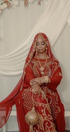 a woman in a red and gold bridal gown sits on a chair with her hands clasped