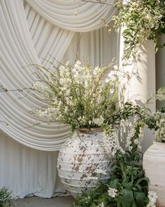 white vases with flowers and greenery in front of a drapeed curtain