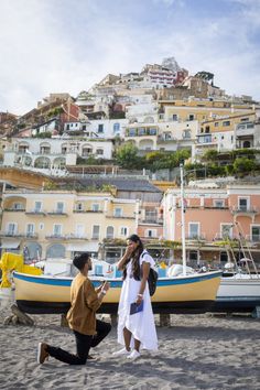 a man kneeling down next to a woman on top of a sandy beach with boats in the background