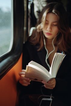 a woman sitting on a train reading a book and listening to headphones with her eyes closed