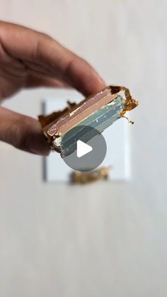 a hand holding a piece of cake with gold and blue icing on it in front of a white wall