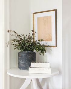 a white table topped with a potted plant next to a framed painting on the wall