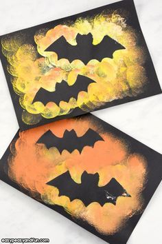 two bats painted on black paper with orange and yellow paint in the background, sitting next to each other