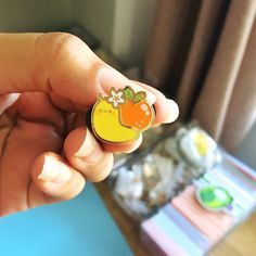 a hand holding a small yellow and orange pin with an apple on it's side