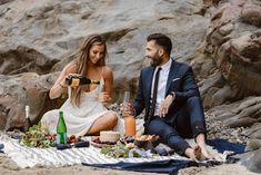 a man and woman sitting on the beach with food, drinks and snacks in front of them