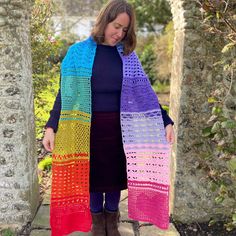 a woman wearing a multicolored crochet scarf standing in an archway with her hands on her hips