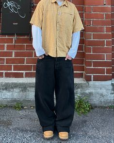 a man standing in front of a brick wall wearing black pants and a yellow shirt