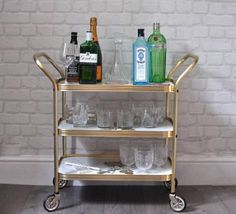 a gold bar cart with glasses and bottles on it next to a white brick wall