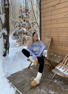 a woman sitting on a wooden bench next to a snow covered forest and holding a coffee cup