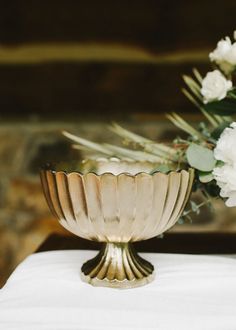 a gold bowl with flowers in it sitting on a white cloth next to a brick wall