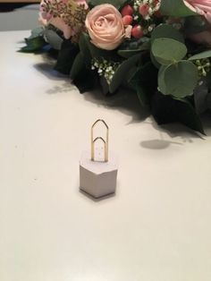 a small white box with a ring on it next to some pink flowers and greenery