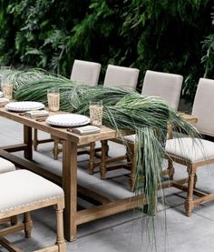 an outdoor dining table with white chairs and green plants on the wall in the background