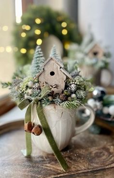 a white mug with a birdhouse in it on top of a table next to a christmas tree