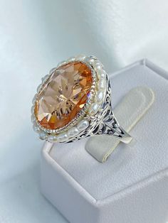 Peach Topaz & Pearl Ring SP (Seed Pearl) Design#3 This is a stunning simulated Peach Topaz gemstone ring. The man-made peach gem has amazing color and perfect clarity. This gemstone is 18mm x 13mm. The garden themed filigree circles the edge of the ring and extends down the band accenting the beautiful large gemstone. Seed pearls surround the large 18 carat gemstone, creating a vibrant contrast. Holding the seed pearls in place are 4 silver leaves edges and fine wire. The hand-strung seed pearls Elegant Peach Colored Ring Jewelry, Elegant Peach Ring Jewelry, Elegant Peach Colored Ring, Elegant Peach Ring, Elegant Peach Pearl Jewelry, Elegant Peach Gemstone Ring, Elegant Peach-colored Ring, Luxury Antique Style Pearl Ring With Cabochon, Peach Gemstone Jewelry