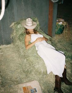 a woman in a white dress and cowboy hat laying on hay