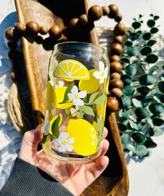 a hand holding a glass with lemons and flowers on it next to a wooden tray