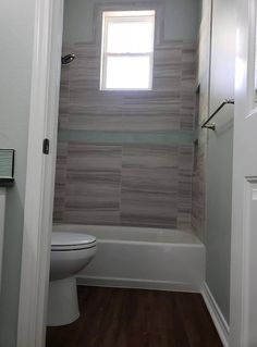 a white toilet sitting under a window next to a bath tub in a bathroom with wooden floors