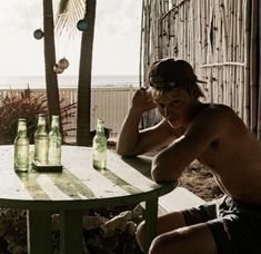 a shirtless man sitting at a table with beer bottles in front of him on the beach