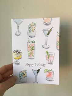 a hand holding up a birthday card with cocktails and drinks in glasses on it