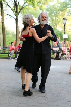 an older man and woman dancing in the park with people sitting on benches behind them