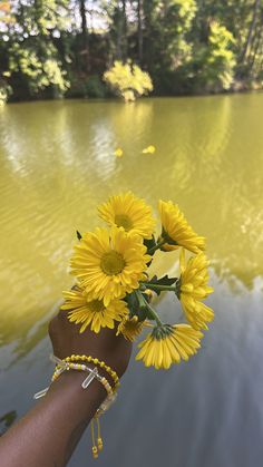 a person's hand holding some yellow flowers near a body of water with trees in the background