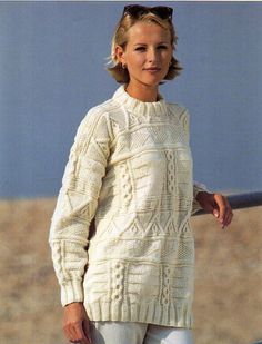 a woman standing on the beach wearing a sweater and white pants with her hands in her pockets