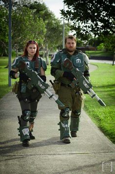 two people dressed in costume walking down a sidewalk with large metal equipment on their shoulders