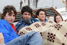 three young men sitting on top of a couch under a blanket next to a body of water