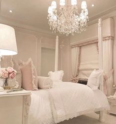 a white bed sitting under a chandelier next to a window with curtains and pillows