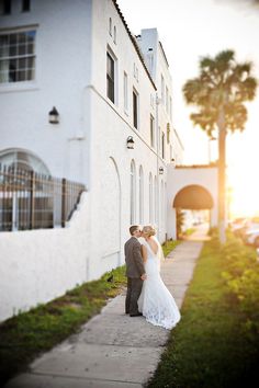 a bride and groom standing on the sidewalk in front of a white building at sunset