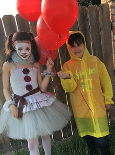 two children dressed up as clowns with balloons in their hands, standing next to a fence