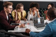 group of people sitting around a table looking at something on the computer screen and laughing