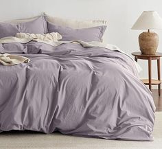 a bed with purple sheets and pillows in a white room next to a night stand